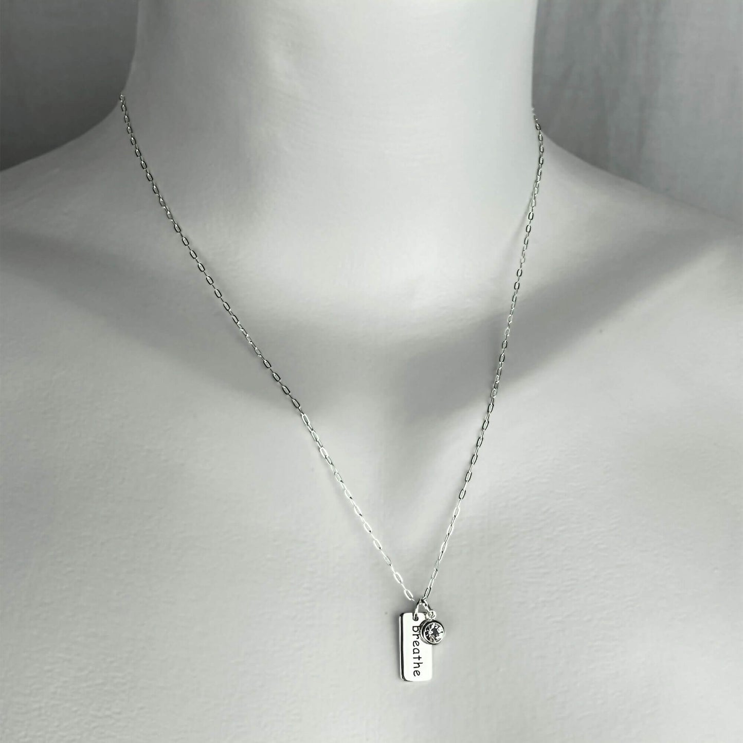 A woman wearing a sterling silver chain necklace with a yoga breathe word small charm accented with a clear crystal dangle charm.
