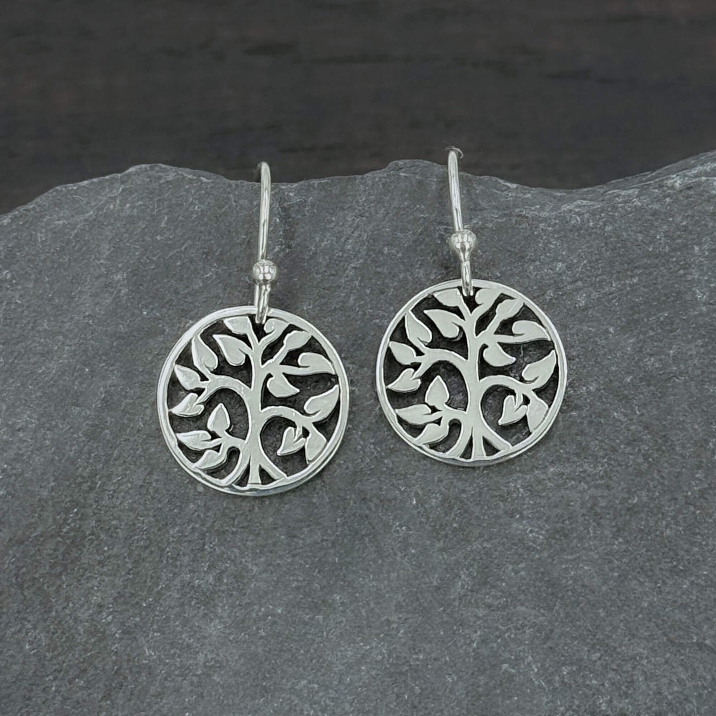 Close up view of 925 solid sterling silver earrings dangling from hook earwires with small circular Tree of Life charms with an openwork cut out design. The tree charms are 5/8" tall and dangle freely. A perfect jewelry gift for nature lovers.