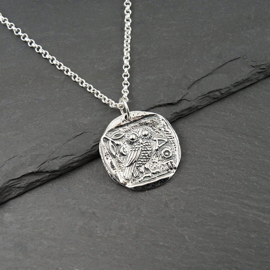 Close-up of a round coin-shaped silver AOE Athena owl pendant on a chain.