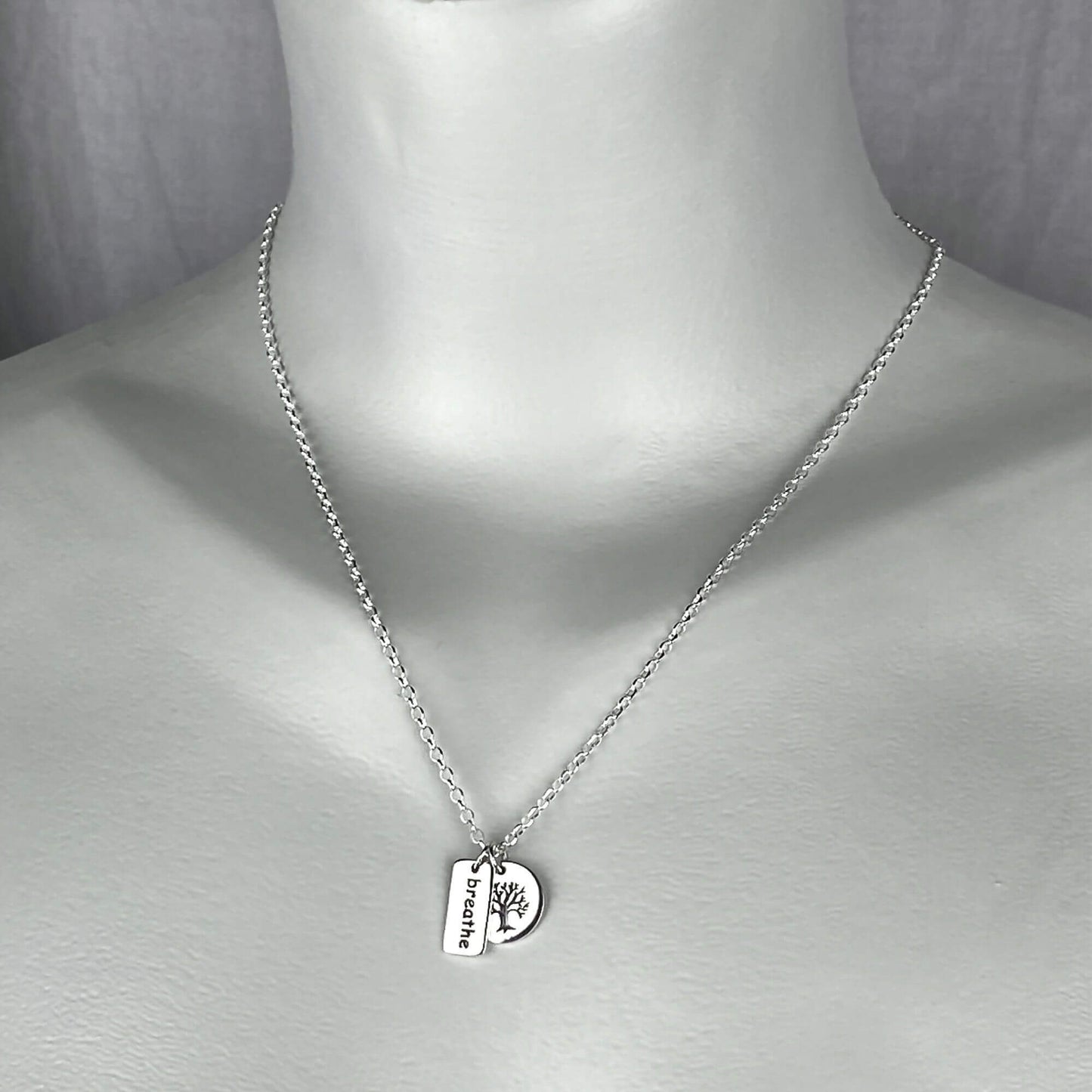 A woman wearing a sterling silver chain necklace with a two small charm: breathe saying with rectangle shape and a round etched tree charm.