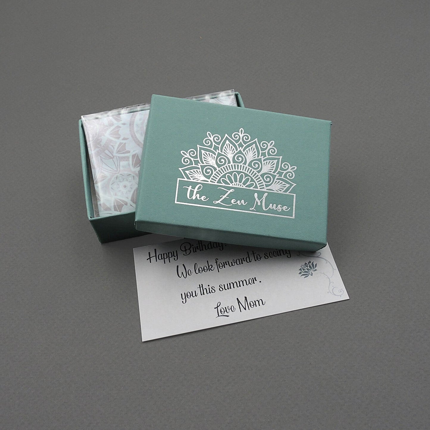 A jewelry gift box with a metallic silver The Zen Muse mandala logo and a matching mandala-patterned note.