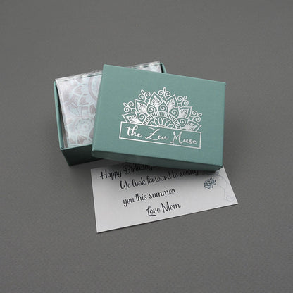 A jewelry gift box with a metallic silver The Zen Muse mandala logo and a matching mandala-patterned note.