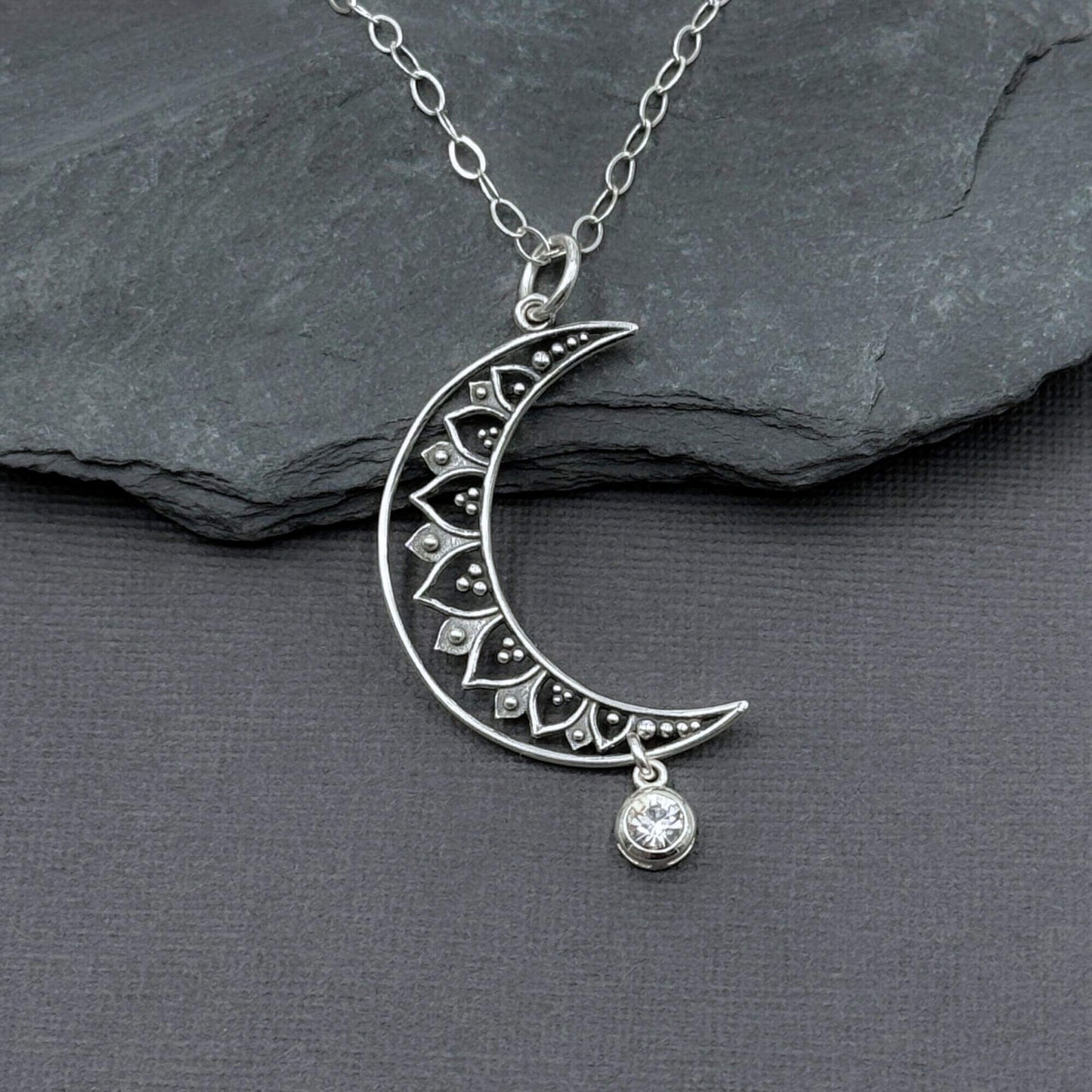 Close-up view of a 925 solid sterling silver crescent moon with cut-out filigree design and a small, clear glass diamond color crystal dangle charm hanging off the bottom tip of the moon.