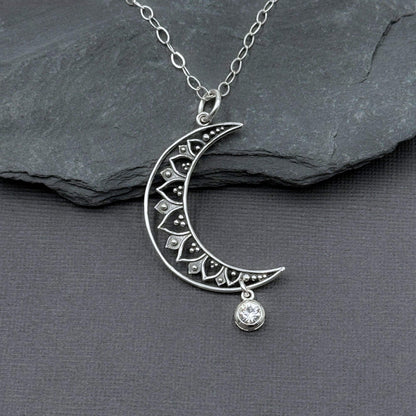 Close-up view of a 925 solid sterling silver crescent moon with cut-out filigree design and a small, clear glass diamond color crystal dangle charm hanging off the bottom tip of the moon.