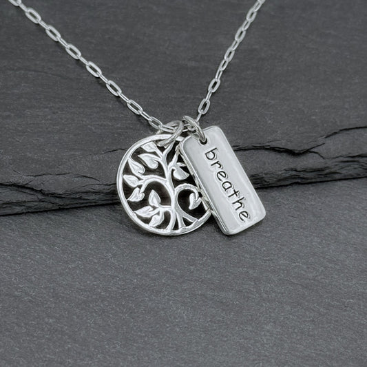 Close-up view of a 925 solid sterling silver charm necklace with two small charms: a round tree of life with an openwork cutout design about 5/8" tall, and a small rectangle charm with the word Breathe etched on it.  The two small charms dangle freely of one another—perfect gift for yoga lovers.